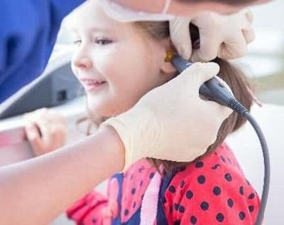 Medicul examinează auzul unei fetițe cu un aparat auditiv special, subliniind importanța diagnosticării timpurii a problemelor de auz la copii.