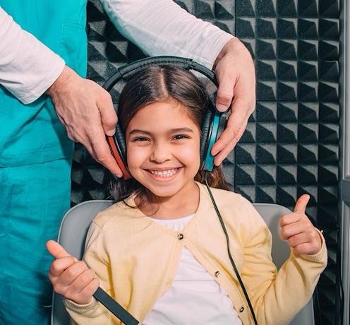 Fetiță zâmbitoare purtând căști în timpul unui test auditiv realizat de medic ORL într-o cameră antifonată – evaluare profesională a auzului la copii.