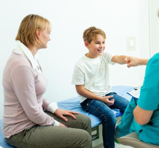Băiat zâmbitor la consult ORL, însoțit de mamă, discutând cu medicul specialist într-un cabinet modern — importanța controalelor periodice pentru sănătatea auzului.