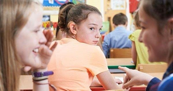 Fetiță cu aparat auditiv privită ironic de alți colegi în clasă — ilustrare a dificultăților sociale și emoționale cauzate de hipoacuzie la copii