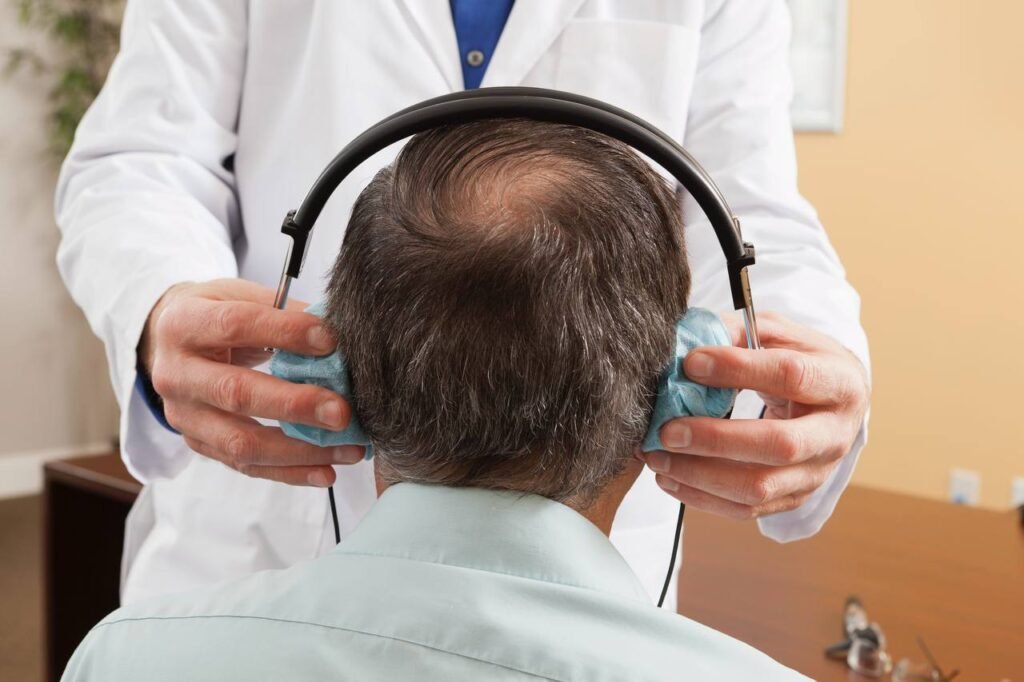 Medic aplică căști speciale unui pacient matur pentru testarea auzului – vedere din spate.