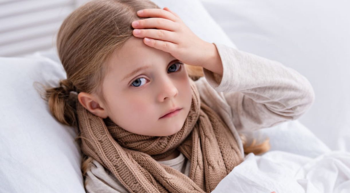 Fetiță care stă în pat și își atinge fruntea, semn de febră sau răceală